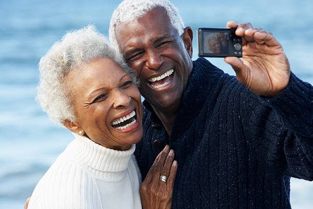 Senior Couple With Camera On Beach Senior Couple With Camera On Beach Taking A Picture Of Themselves Laughing old black couple in love stock pictures, royalty-free photos & images