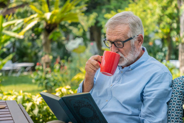 Senior elderly man reading book drinking mug of coffee in garden stock photo
