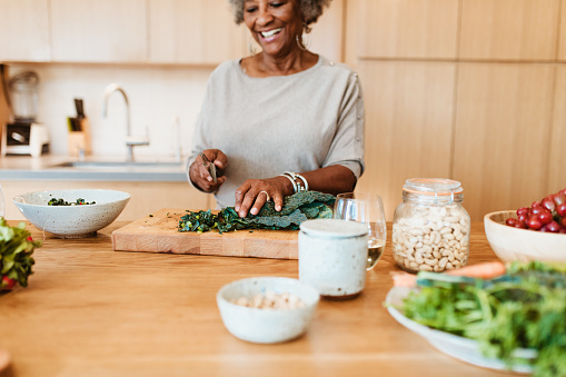 Smiling retired woman preparing meal in kitchen. Senior female is chopping vegetable at kitchen island. She is having frizzy gray hair.