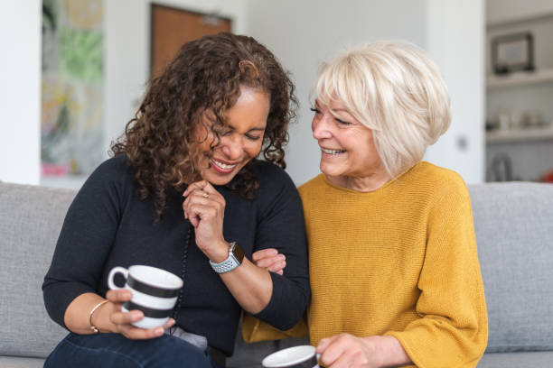 Senior female friends having tea Two senior women are spending time together. The friends are seated next to each other indoors. They are holding cups of coffee. One woman is caucasian and the other woman is black. The friends are embracing and laughing. baby boomers stock pictures, royalty-free photos & images