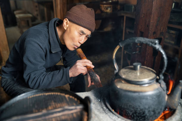 senior japanese man blowing through bamboo to add oxygen to fire in wood burning stove in kitchen - idosos aquecedor imagens e fotografias de stock