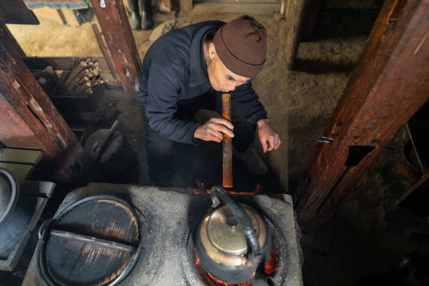 senior japanese man blowing through bamboo to add oxygen to fire in wood burning stove in kitchen - idosos aquecedor imagens e fotografias de stock