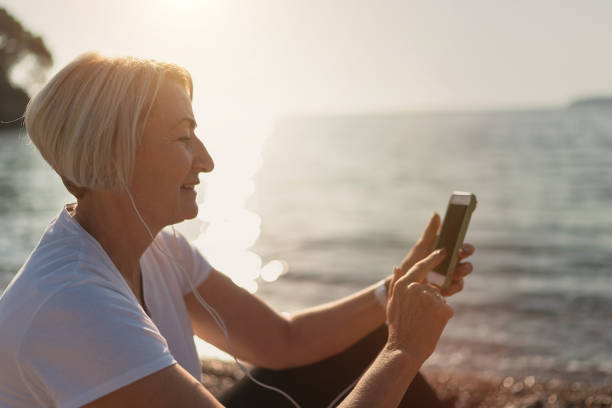 senior dam med smartphone och hörlurar vid solnedgången - senior listening music beach bildbanksfoton och bilder