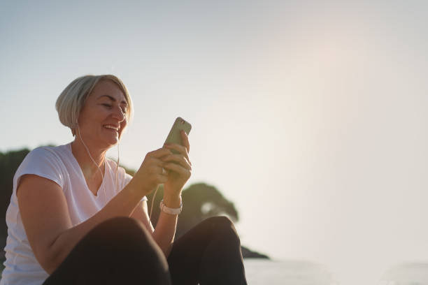 senior dam med smartphone och hörlurar vid solnedgången - senior listening music beach bildbanksfoton och bilder