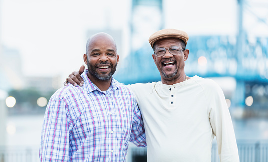 A senior African-American man in his 60s and his son-in-law, in his 40s, standing side by side outdoors on a city waterfront, a bridge out of focus in the background. They are posing for the camera, arms around each other, smiling.