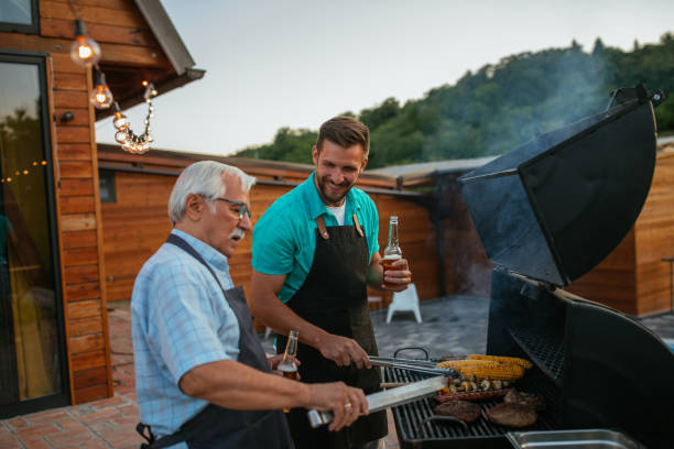 Senior man and his son roasting meat on the BBQ grill in the backyard and drinking beers Pensioner and his son wearing aprons, holding bottles of beer and roasting meat on barbecue in the backyard fathers day stock pictures, royalty-free photos & images