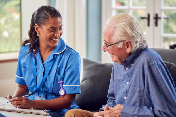 Senior Man At Home Talking To Female Nurse Or Care Worker In Uniform Making Notes In Folder stock photo