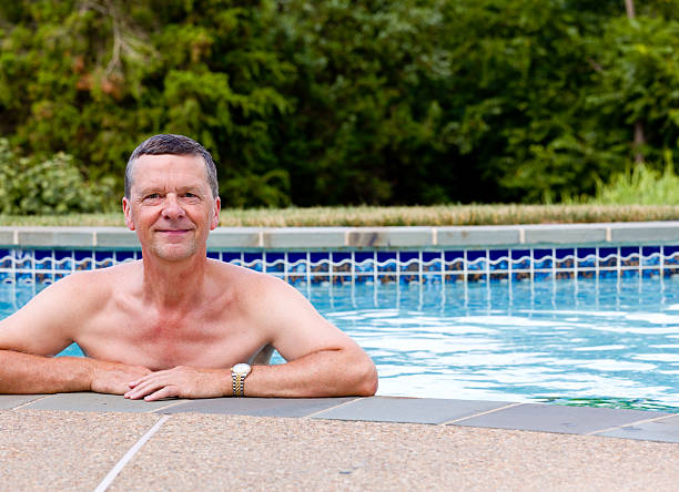 Senior male relaxing by the side of a modern swimming pool in back...