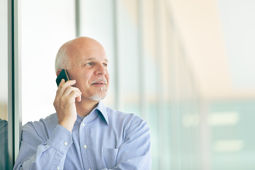 Senior businessman chatting on his smartphone in a high key portrait as he stands leaning against glass windows of a commercial building or office block looking to the side with a quiet smile