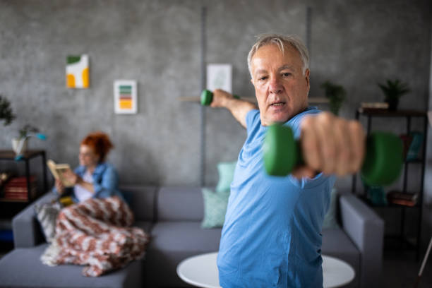 Senior man exercising with hand weights while his wife relaxing on sofa and reading book stock photo