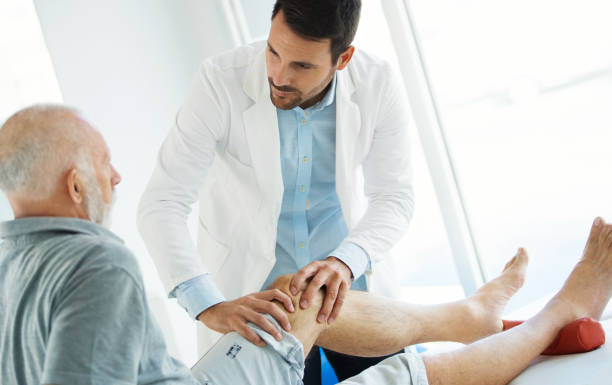 Senior man having his knee examined by a doctor. Closeup over the shoulder view of an early 60's senior gentleman having some knee pain. He's at doctor's office having medical examination by a male doctor. The doctor is touching the sensitive area and trying to determine the cause of pain. osteoarthritis stock pictures, royalty-free photos & images