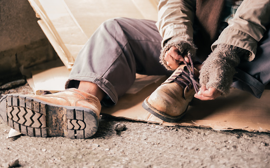 Senior man homeless wearing sweater and cloth gloves with sitting on cardboard and tying laces on shoes. Close up of asian man homeless in shelter and wear leather shoes boot and tie lace his shoes.