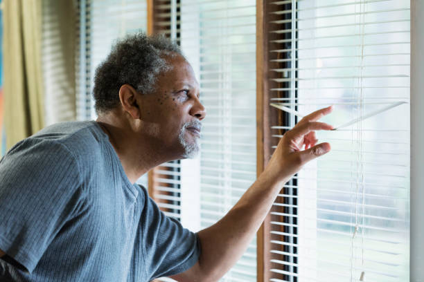 Senior man looking out the window An African-American senior man in his 60s at home, looking out the window through the blinds with a serious expression on his face. sad old black man stock pictures, royalty-free photos & images