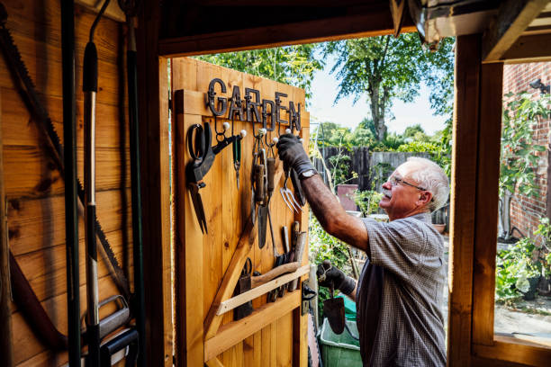 Senior man selecting hand tool from door of gardening shed View from inside storage shed as Caucasian man in late 60s wearing casual clothing and work gloves selects tool from hook on door. shed stock pictures, royalty-free photos & images