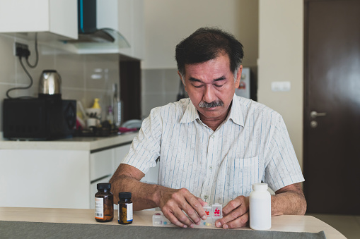 Elderly Asian man sort his weekly prescription medication and putting them into a daily pill organizer box on desk. Home Medical & Healthcare, Telehealth & Telemedicine, Stay at Home Concepts.