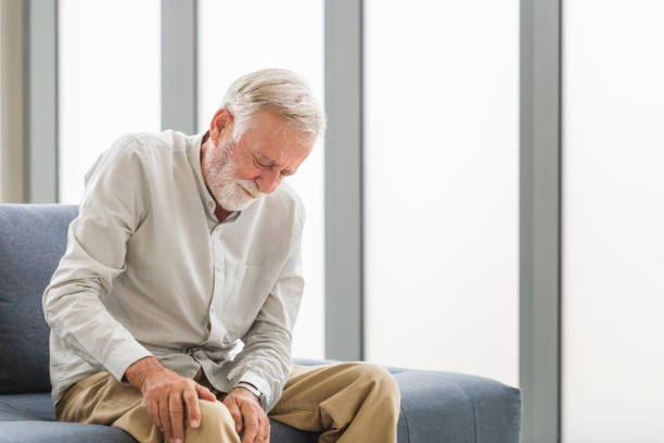 Senior man suffering from knee pain sitting sofa in the living room, Elderly man suffering from knee pain while sitting on the sofa stock photo