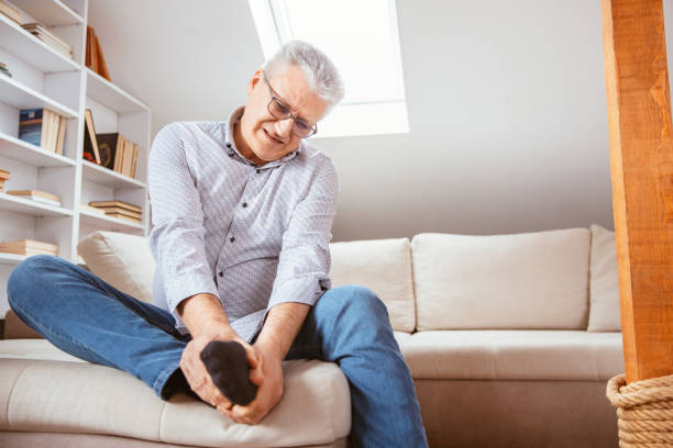 Senior man suffering with foot cramp Senior man suffering with foot cramp on sofa in living room at home. foot stock pictures, royalty-free photos & images