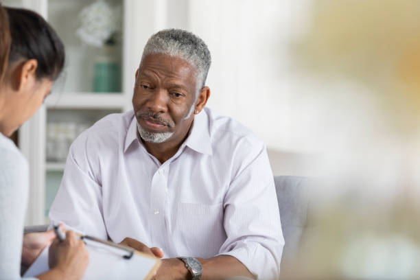 Senior man talks to therapist as she takes notes A senior man sits on a couch opposite his unrecognizable female therapist and looks down with a sad expression.  His therapist takes notes with a pen and clipboard. sad old black man stock pictures, royalty-free photos & images