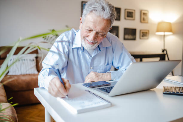 Senior man using a laptop at the table and writing notes stock photo