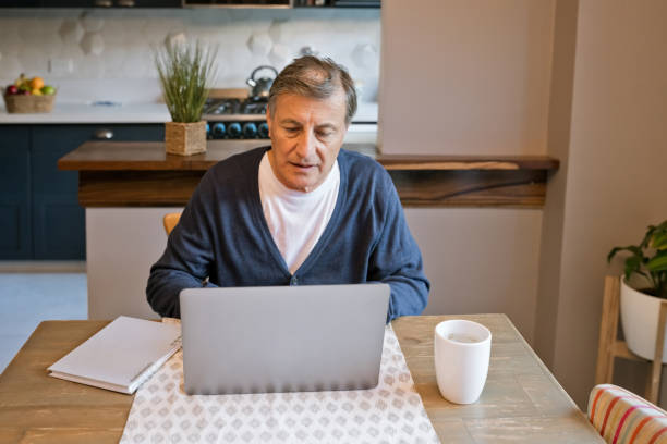 Senior man using laptop at home Elderly male sitting at the table at home and using laptop. Active senior working on computer. social work online stock pictures, royalty-free photos & images