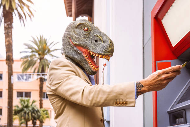 戴著 t-rex 恐龍面具的高級男子用轉帳卡從銀行收銀機取款--一半人和動物的超現實形象--atm 的荒謬和瘋狂的自動取款機廣告概念 - england australia 個照片及圖片檔