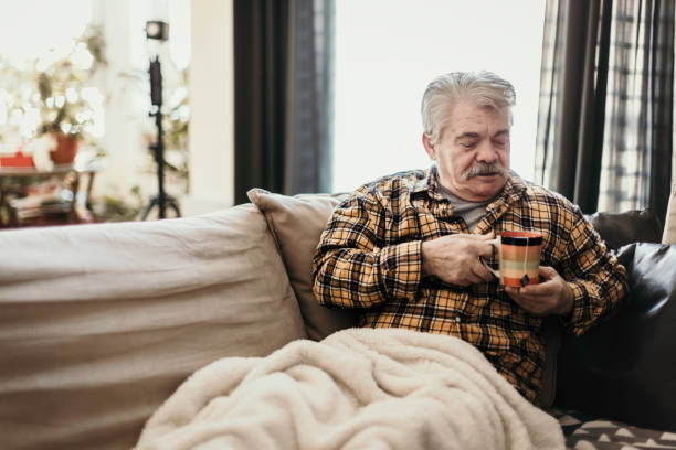 Senior man with flu sitting on sofa wrapped in a warm blanket stock photo