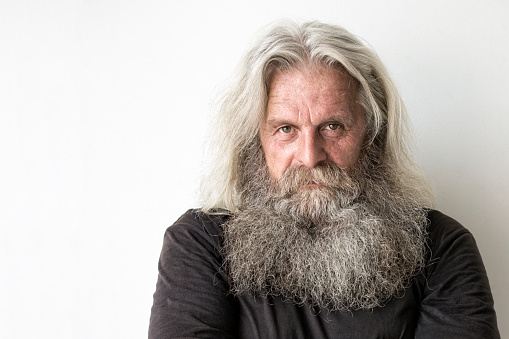 A close-up portrait of a senior man with long grey hair and a long beard standing in front of a white wall. He is wearing a black shirt and he is looking straight and serious at the camera. There is some copy space on the left side of the photo.