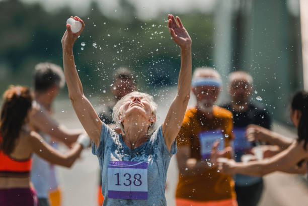 Senior marathon runner refreshing herself with water during a race. Senior woman running a marathon on the road and refreshing herself with water from a cup. There are people in the background. marathon stock pictures, royalty-free photos & images