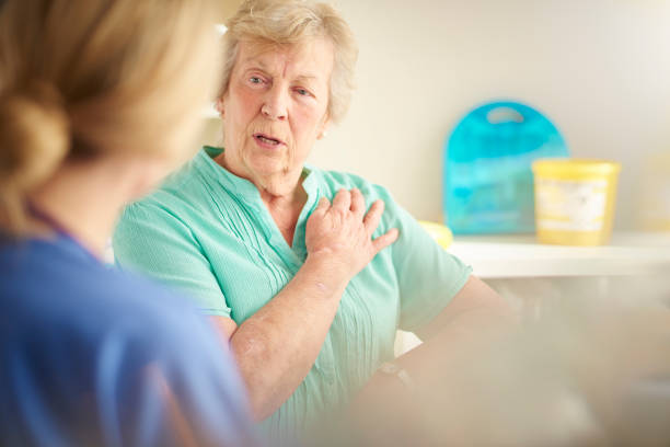 Senior patient complaining of chest pain a senior female patient sits and chats to a female doctor who we see from behind . She is listening to her symptoms chest pain stock pictures, royalty-free photos & images