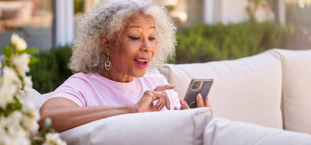 Senior Retired Woman Sitting Outside In Garden At Home Making Video Call On Mobile Phone stock photo
