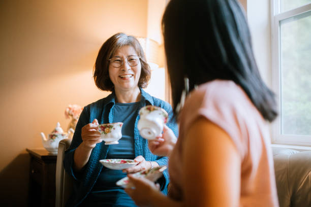 Senior Woman and Her Daughter Having Tea Together A loving Korean mother in her 60's enjoys time with her adult daughter, drinking a cup of tea on the living room sofa. asian mother talking with daughter stock pictures, royalty-free photos & images