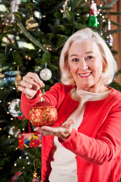 senior woman decorating christmas tree holding ornament - vrouw kerstboom versieren stockfoto's en -beelden