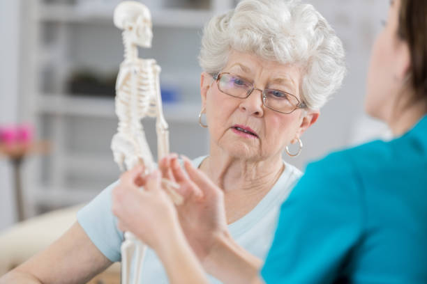 Senior woman discusses posture with physical therapist Senior woman listens seriously as her physical therapist sits with her and holds a model of the human skeletal system.  The therapist is discussing the affects of osteoporosis in posture and the reason for resulting pain. osteoporosis photos stock pictures, royalty-free photos & images