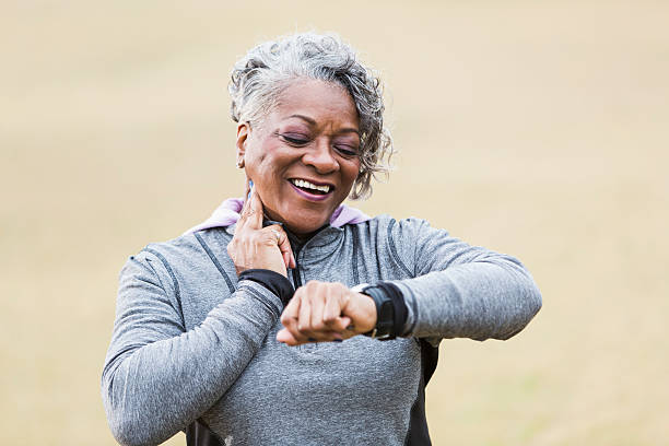 An African American senior woman, in her 60s, exercising outdoors in the park.  She is checking her pulse to see if her exercise has increased her heart rate.  She is smiling, with two fingers on her neck, looking at the watch on her wrist.  She is wearing a gray sweatshirt.