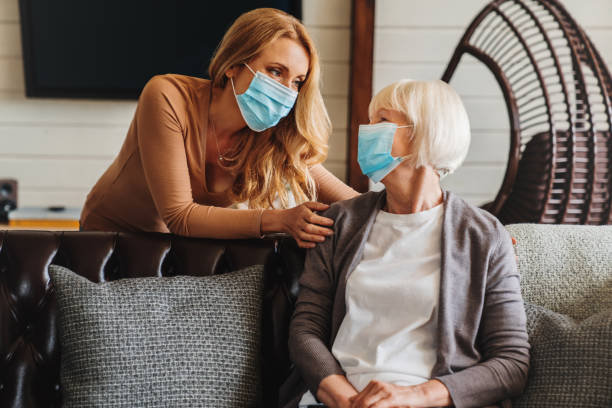 seniorin in medizinischer maske mit sozialarbeiterin zu besuch zu hause - krankheit fotos stock-fotos und bilder