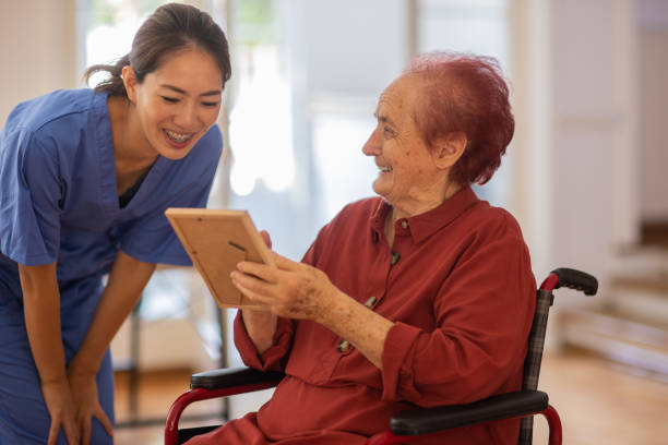 Senior woman in wheelchair talking funny things from life while showing her photo to nurse at nursing home stock photo