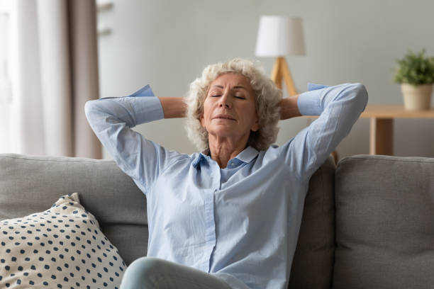 Senior woman putting hands behind head enjoying fresh air Old 70s woman dreamer seated on couch relaxing meditating thinking positive thoughts putting hands behind head closed eyes breathing fresh air relish retired life free time at home, well-being concept deep stock pictures, royalty-free photos & images