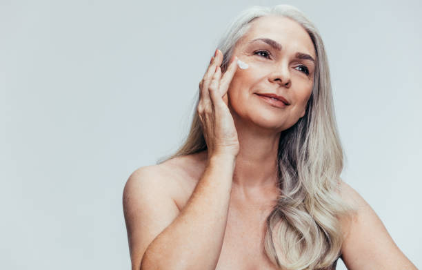 Senior woman putting on anti aging cream Senior woman putting on anti aging cream on her face and looking away. Female applying moisturizer lotion on her face against grey background. applying face cream stock pictures, royalty-free photos & images