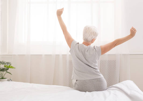 Senior woman sitting on her bed in morning Senior woman sitting on her bed in morning, stretching with arms raised, back view, free space waking up stock pictures, royalty-free photos & images