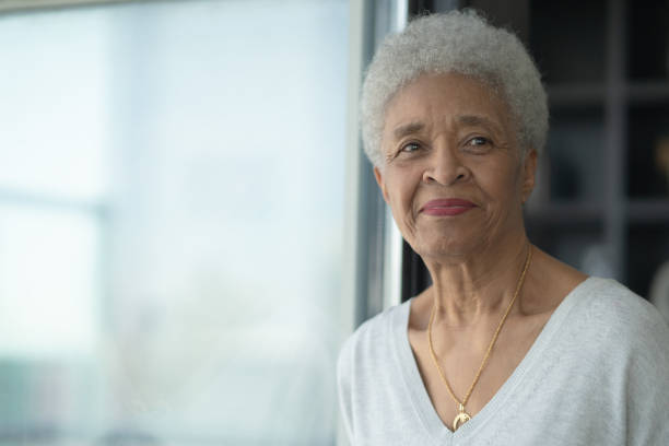 A Senior Woman Smiling As She Reflects stock photo A senior African woman sits beside a window and gazes off into the room as she is deep in thought and reflection.  She has a gentle smile on her face and is wearing a casual and comfortable sweater in this head shot. 50 59 years photos stock pictures, royalty-free photos & images
