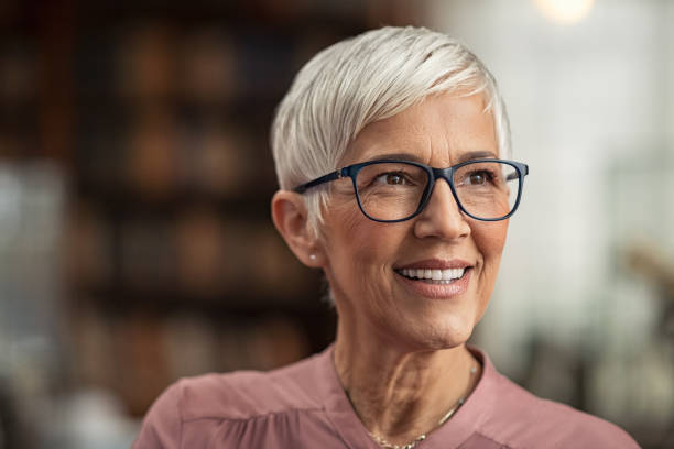 Senior woman smiling with eyeglasses Portrait of smiling mature woman with spectacles and short grey hair in library looking away. Senior librarian standing in reading hall and thinking. Old beautiful lecturer contemplating in library. Future and vision concept. 50 54 years stock pictures, royalty-free photos & images