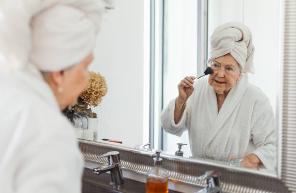 Senior woman standing in front of mirror in her bathroom and preparing her face with make up brushes. stock photo