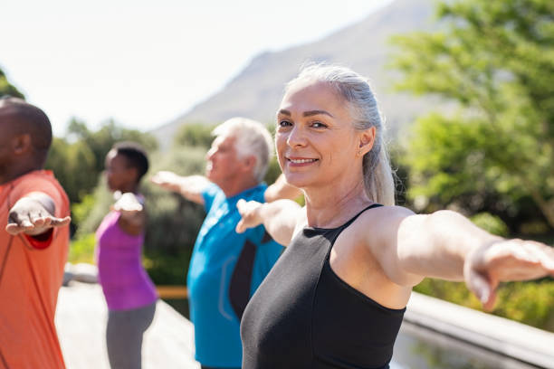 Senior woman stretching arms in yoga class Portrait of happy senior woman practicing yoga outdoor with fitness class. Beautiful mature woman stretching her arms and looking at camera outdoor. Portrait of smiling serene lady with outstretched arms at park. active seniors stock pictures, royalty-free photos & images