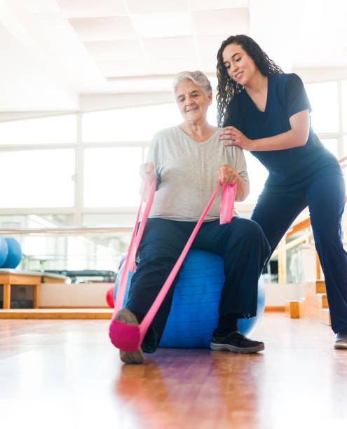 Senior woman stretching leg with exercise band A senior latin woman sitting on a fitness ball, stretching her leg with an exercise band and being helped by the pysical therapist. yoga ball work stock pictures, royalty-free photos & images