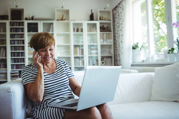 Senior woman talking on mobile phone while using laptop in living room Senior woman talking on mobile phone while using laptop in living room at home irish women stock pictures, royalty-free photos & images