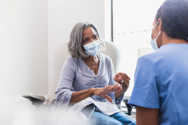 Senior woman talks with female healthcare professional A senior woman, wearing a protective face mask, talks with a female nurse during a medical appointment. woman talking to doctor stock pictures, royalty-free photos & images