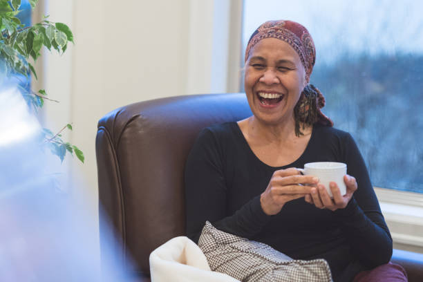 Senior woman with cancer sits by her window drinking tea A beautiful senior woman sits by her living room window and drinks a cup of tea. She is laughing at something one of her friends (off-camera) just said. She is wearing a headscarf. big smile emoji stock pictures, royalty-free photos & images
