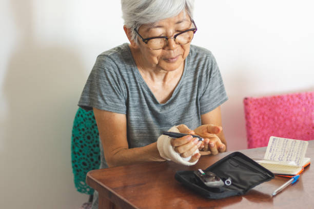 Senior woman with glucometer checking blood sugar level at home Senior woman with glucometer checking blood sugar level at home. She is asian, has white short hair, wears glasses and a bandage on one hand. Medicine, age, diabetes and health care concept hyperglycemia stock pictures, royalty-free photos & images