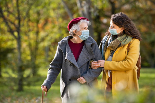 Senior woman with lovely girl wearing face mask at park Lovely granddaughter walking with senior woman holding stick in park and wearing mask for safety against covid-19. Happy old grandmother enjoying walking in park with girl. Smiling elderly woman with happy caregiver in park relaxing after quarantine due to coronavirus outbreak and lockdown. senior adult stock pictures, royalty-free photos & images