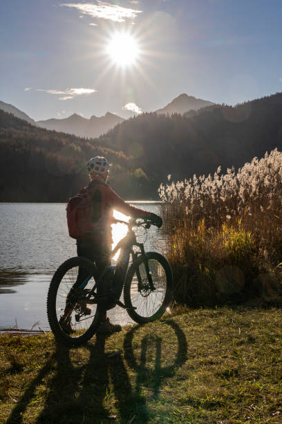 senior woman with mountain bike in bright backlit at lake senior woman with electric mountain bike in bright backlit sun on the shore of lake Weissensee near city of Fuessen, eastern Allgaeu, Bavarian alps allgau alps stock pictures, royalty-free photos & images